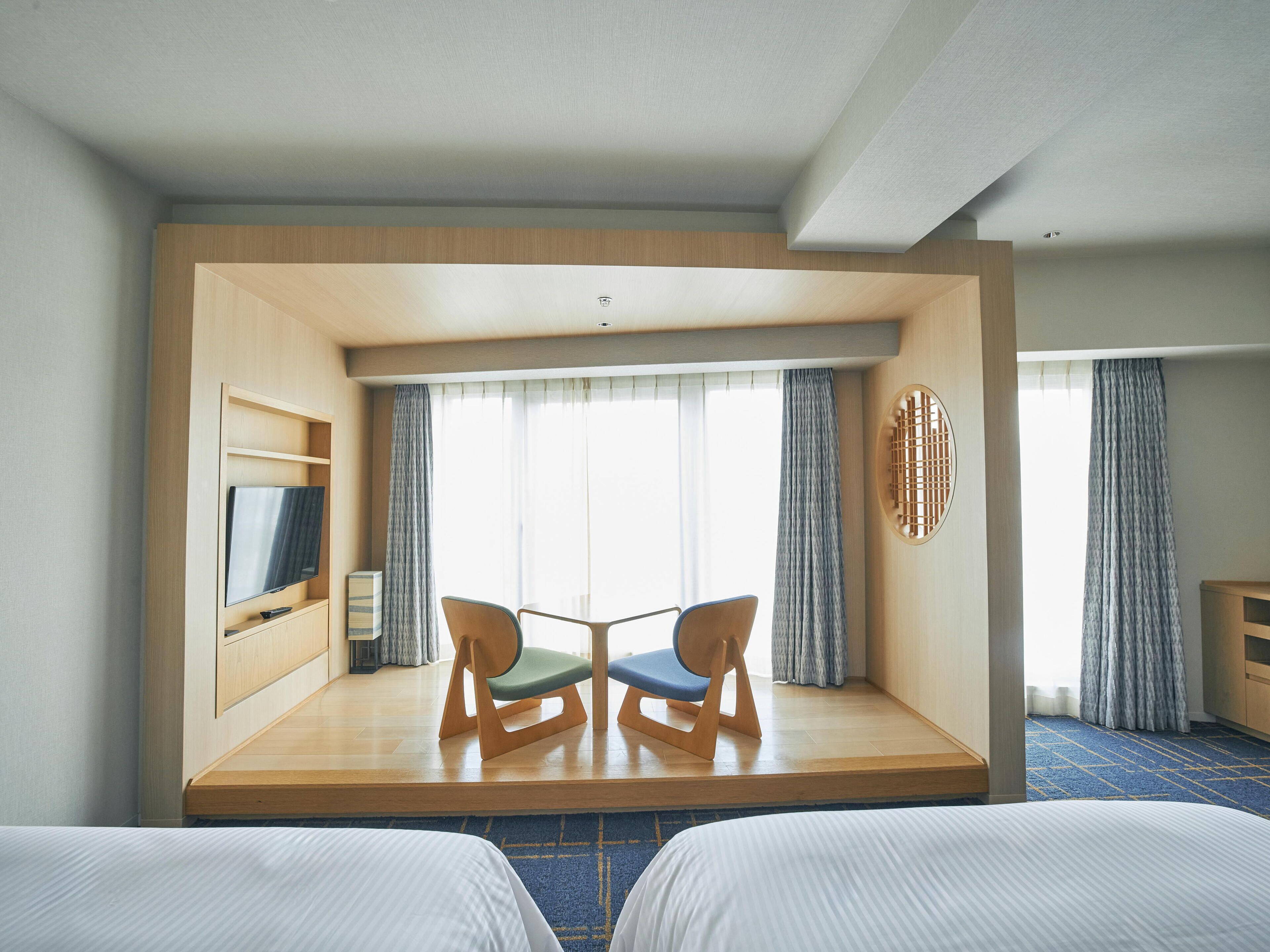 A hotel room with two beds, a TV on a wooden shelf, a small table with chairs, and a large window with curtains.