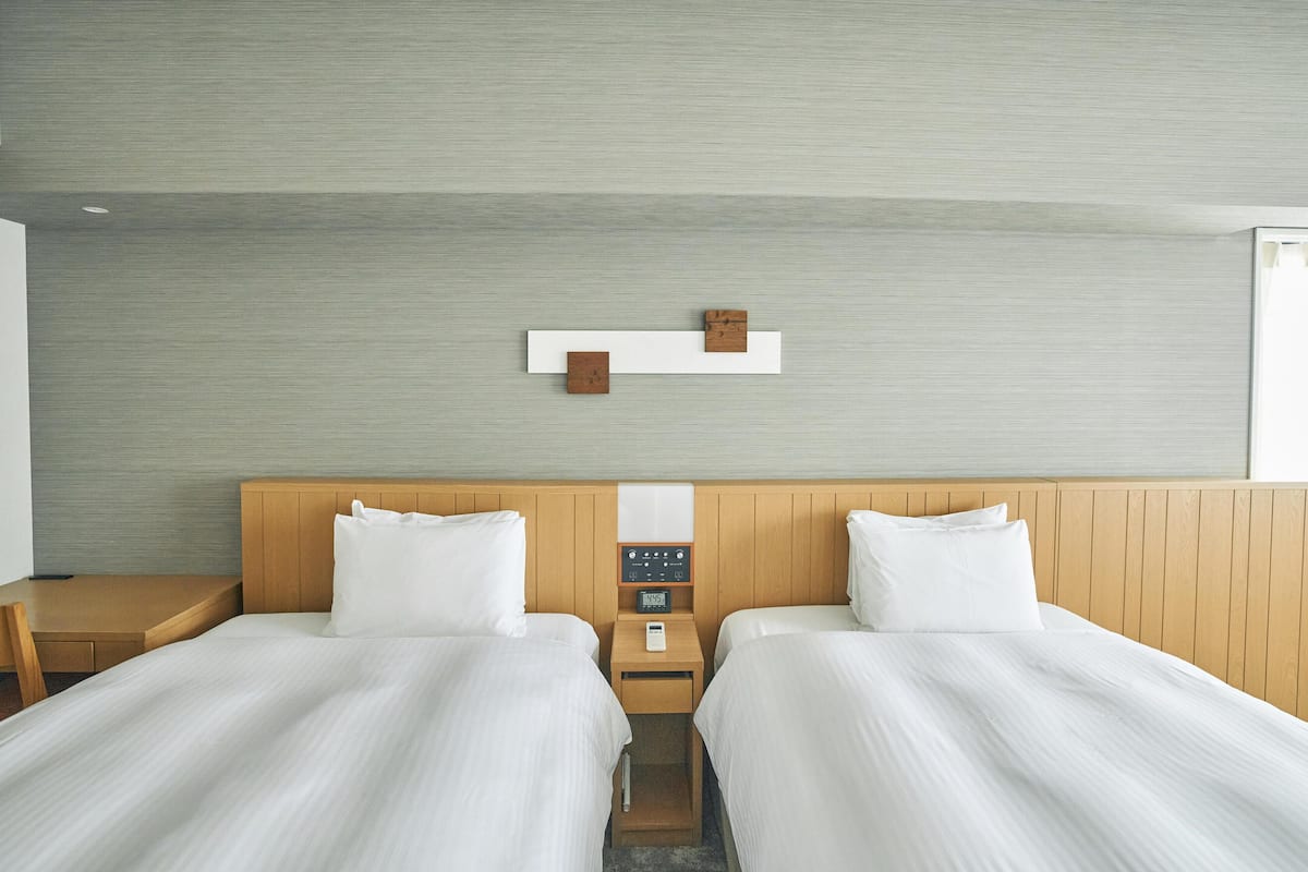 A hotel room with two beds, a wooden headboard, a small bedside table with a clock, and a wall-mounted shelf with a wooden block.