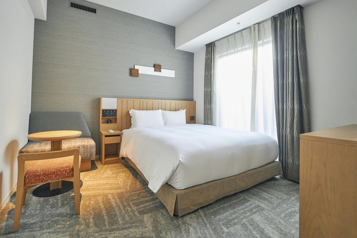 A modern hotel room with a large bed, a desk, a chair, and a wardrobe.