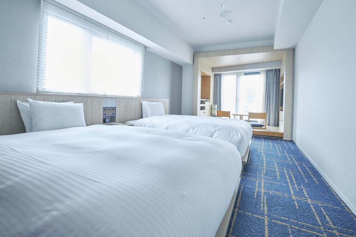 A hotel room with two beds, a large window, a bathroom, and a view of the city.
