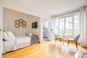 A modern bedroom with a bed, a small table, and a chair. There is a window with curtains, and a view of the outside.