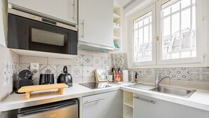 A modern kitchen with a built-in oven, microwave, and dishwasher. It features a wooden cutting board, a kettle, and a sink with a window above it. - Palais des Congres - Studio (Paris)