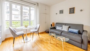 A modern living room with a grey sofa, two armchairs, a glass coffee table, and a round table. There are two windows with white curtains, and a view of buildings outside. - Palais des Congres - Studio (Paris)