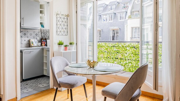 A modern kitchen with a built-in dishwasher, a dining area with a round table and two chairs, and a large sliding glass door leading to a balcony with a green leafy wall. - Palais des Congres - Studio (Paris)