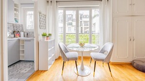 A modern kitchen with white cabinets, a dining area with a round table and two chairs, and a large window with a view of buildings. - Palais des Congres - Studio (Paris)