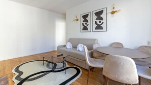Ein modernes Wohnzimmer mit einer Couch, einem runden Tisch und Stühlen. Zwei Wandleuchten und ein abstraktes Kunstwerk sind an der Wand angebracht.