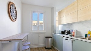 Eine kleine Küche mit Holztisch und Hockern, ein Fenster mit Blick und ein Spülbecken mit verschiedenen Gegenständen auf der Arbeitsfläche.