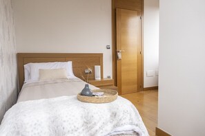Ein ordentlich bezogenes Bett mit weißer Daunendecke, einer grauen Vase und einem gewebten Korb.