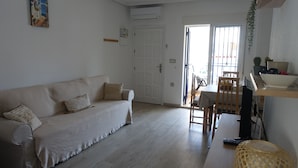 A living room with a beige sofa, a wooden dining table with chairs, and a small kitchen area with a sink and countertop.