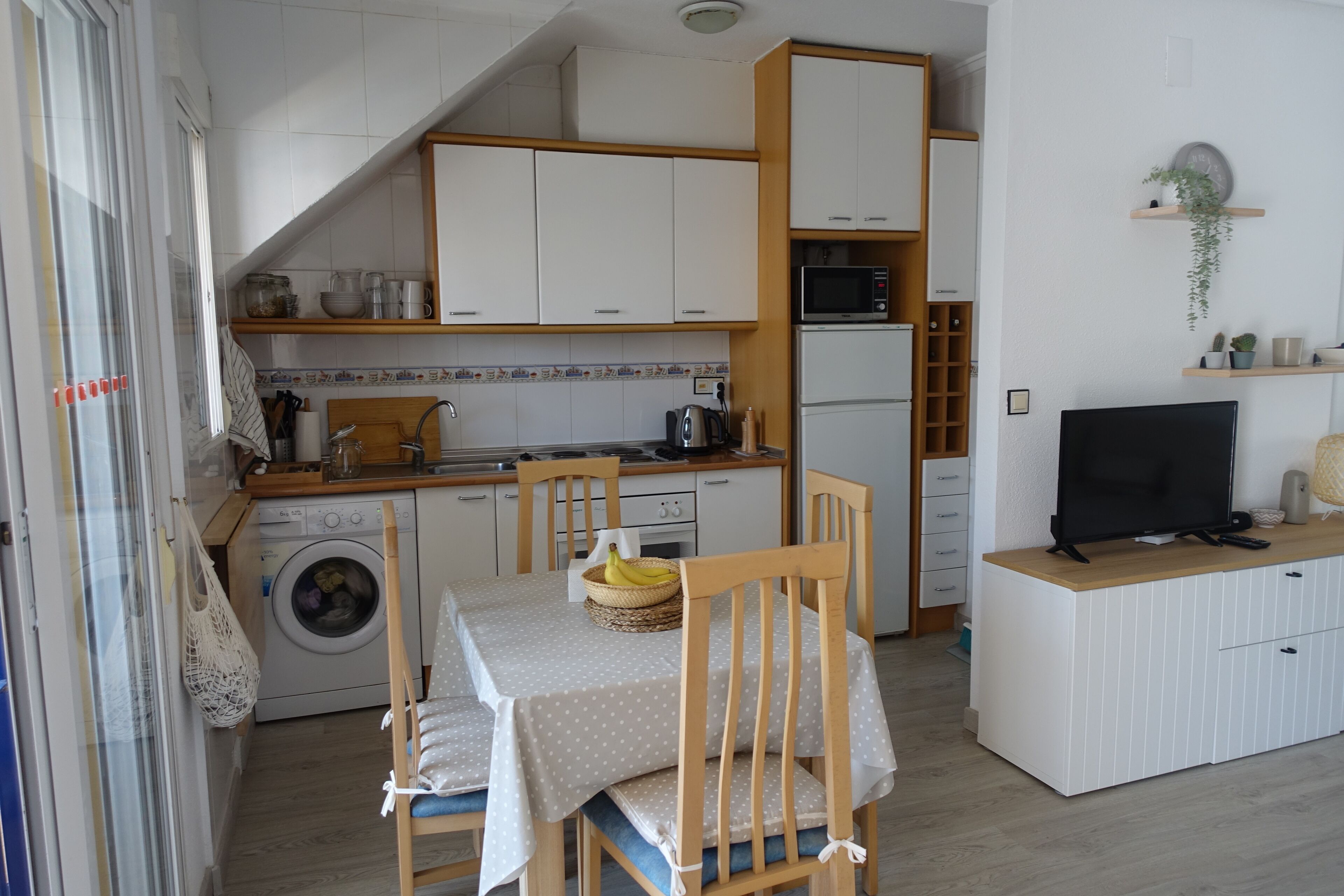 A compact kitchen with a washing machine, a dining table with chairs, a microwave, and a television on a wooden stand.