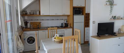 A compact kitchen with a washing machine, a dining table with chairs, a microwave, and a television on a wooden stand.