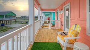 A wooden deck with a green artificial turf, a white chair with an orange cushion, and a pink building with a sign that reads "Ice Cream Only". - Sunset Balcony • Covered Patio Lounge + Mins to Beach | Surf-n-dipity (Port Aransas)