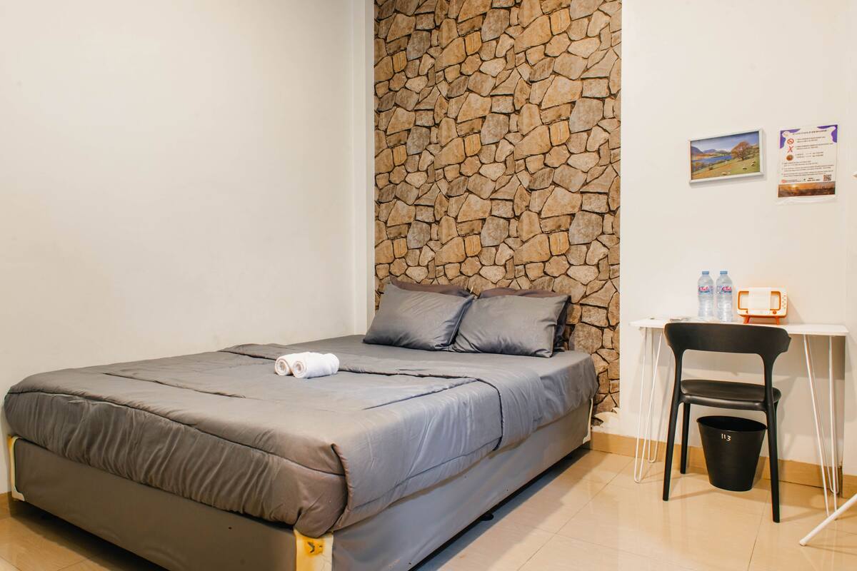 Een bed met grijze beddengoed, een stenen muur, een bureau met stoel en een prullenbak.