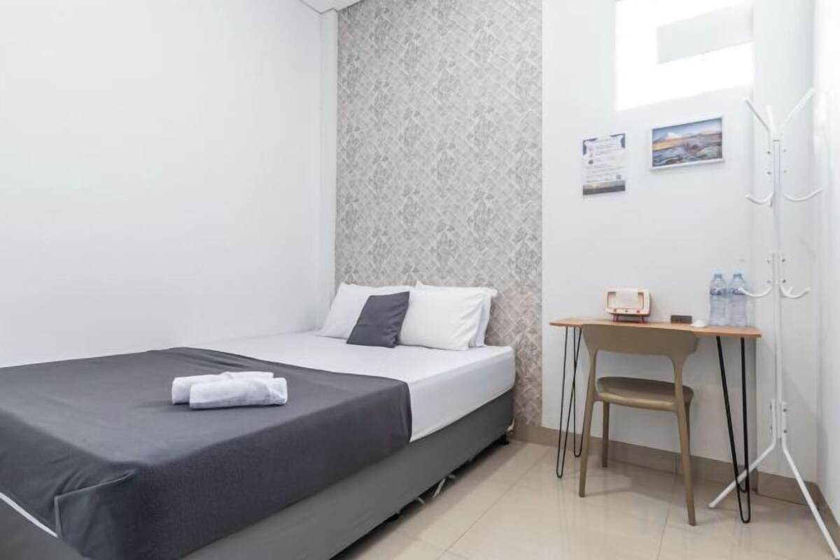 Een enkel bed met een grijze spreidek, een klein houten tafeltje met een stoel, en een haak op de muur.