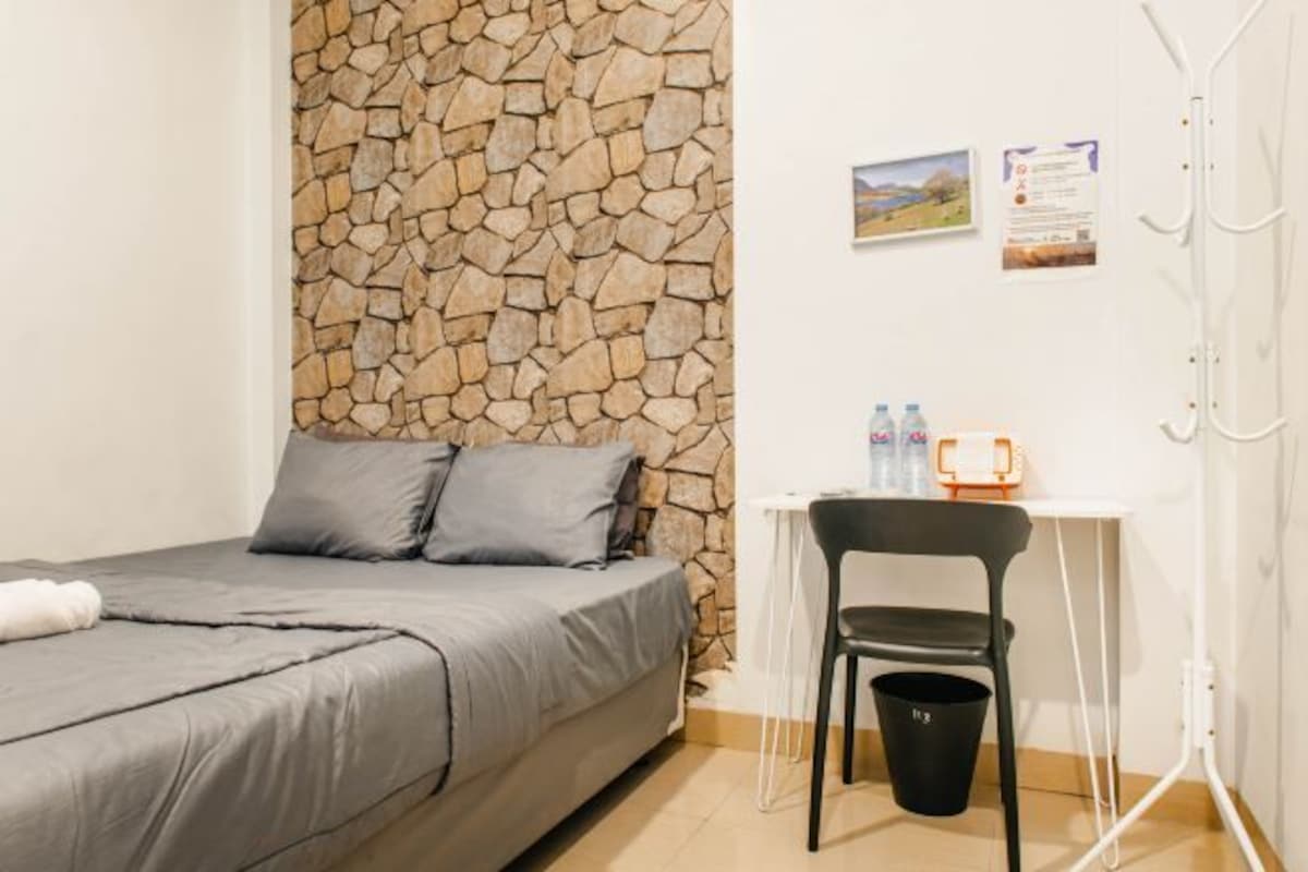 Une chambre avec un mur en pierre, un lit avec une literie grise, un petit bureau avec une chaise et une poubelle.