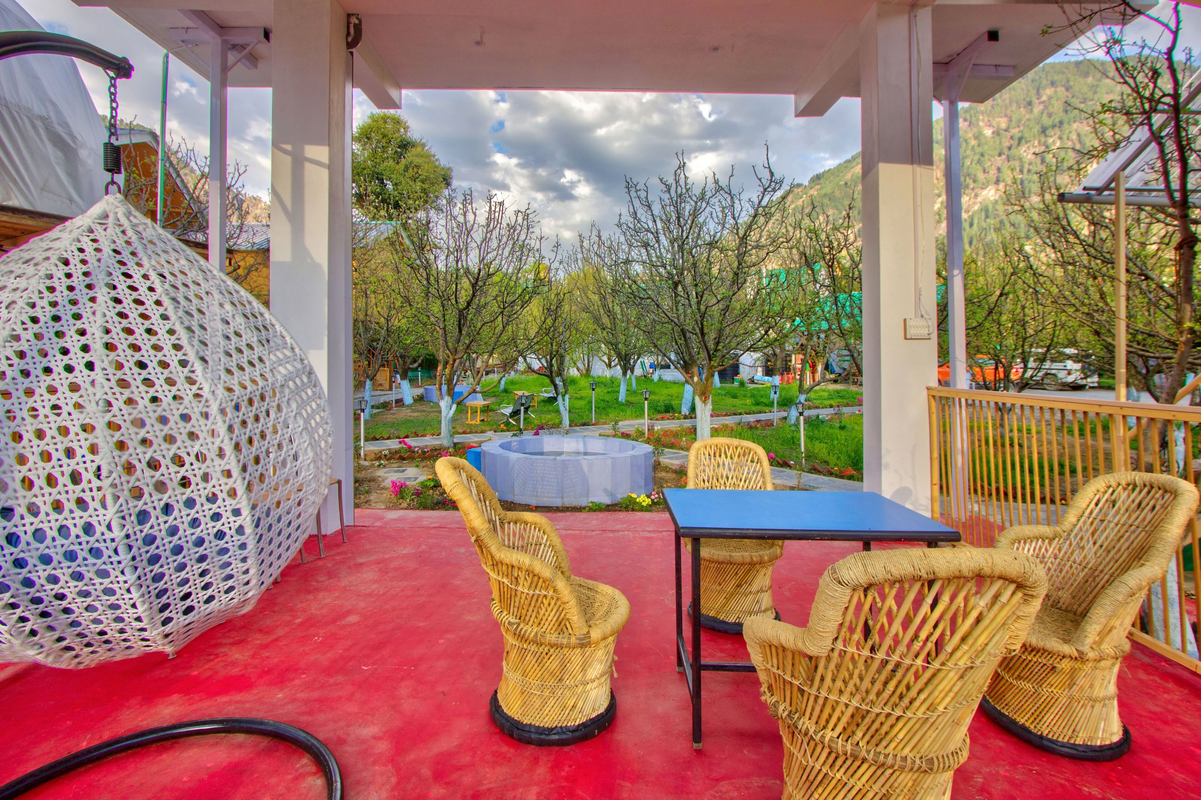 A covered outdoor seating area with wicker chairs and a table, a hanging white crochet structure, and a view of a garden and mountains in the background.