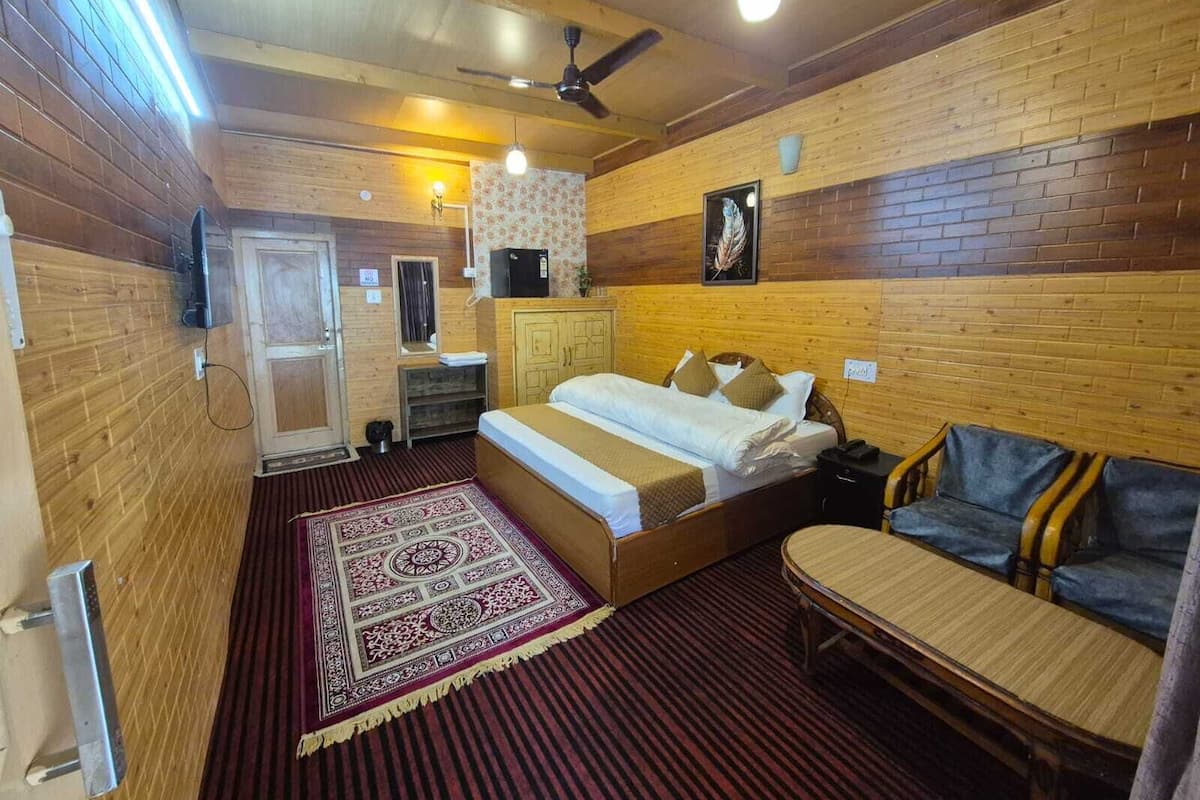 A wooden-paneled room with a bed, a small table, a sofa, a ceiling fan, and a wall-mounted TV.