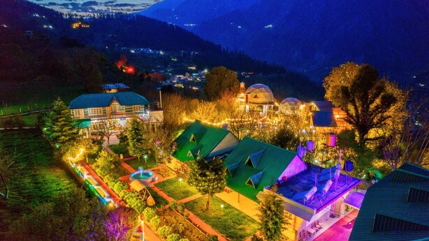 Un hotel con varias construcciones, incluyendo una edificación principal con techo verde y otras más pequeñas. Se observan árboles y montañas al fondo.