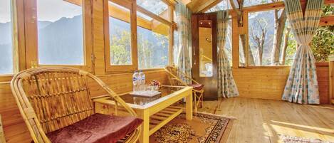 Eine Holzhütte mit großen Fenstern, einem Korbstuhl, einem Holztisch und einem gemusterten Teppich.
