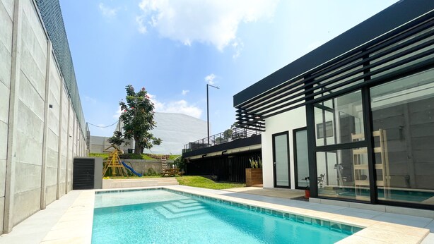 Un área moderna de piscina al aire libre con una piscina azul cristalina, junto a un edificio con ventanas amplias y un balcón.