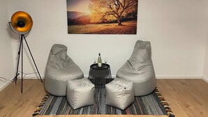 Et værelse med to grå puf, et lille rundt bord med en flaske og glas, en gulvlampe og et billede af et træ i ramme på væggen.