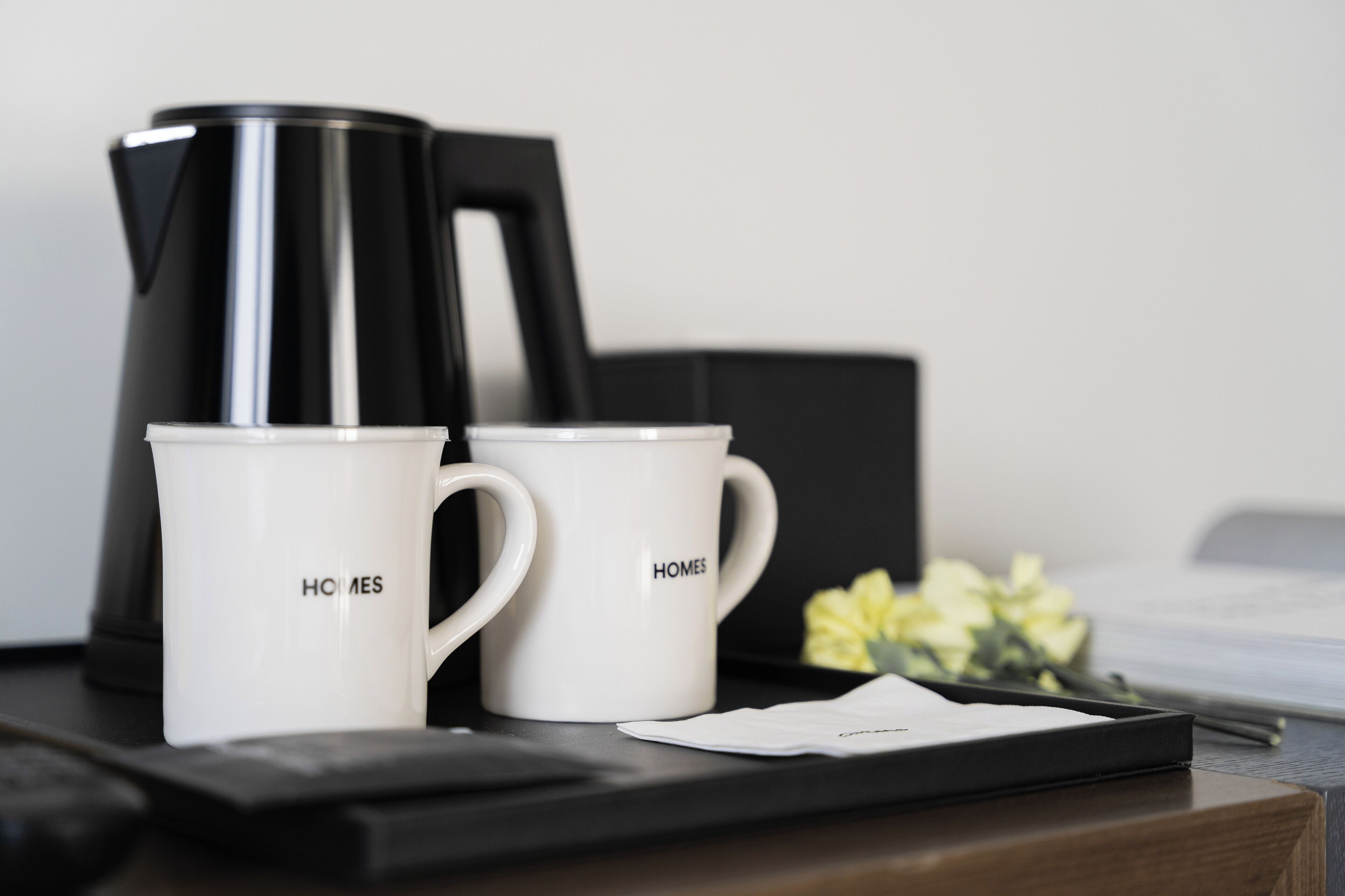 Two white mugs with "HOMES" printed on them, a black coffee maker, and a tray with a napkin on a wooden surface.