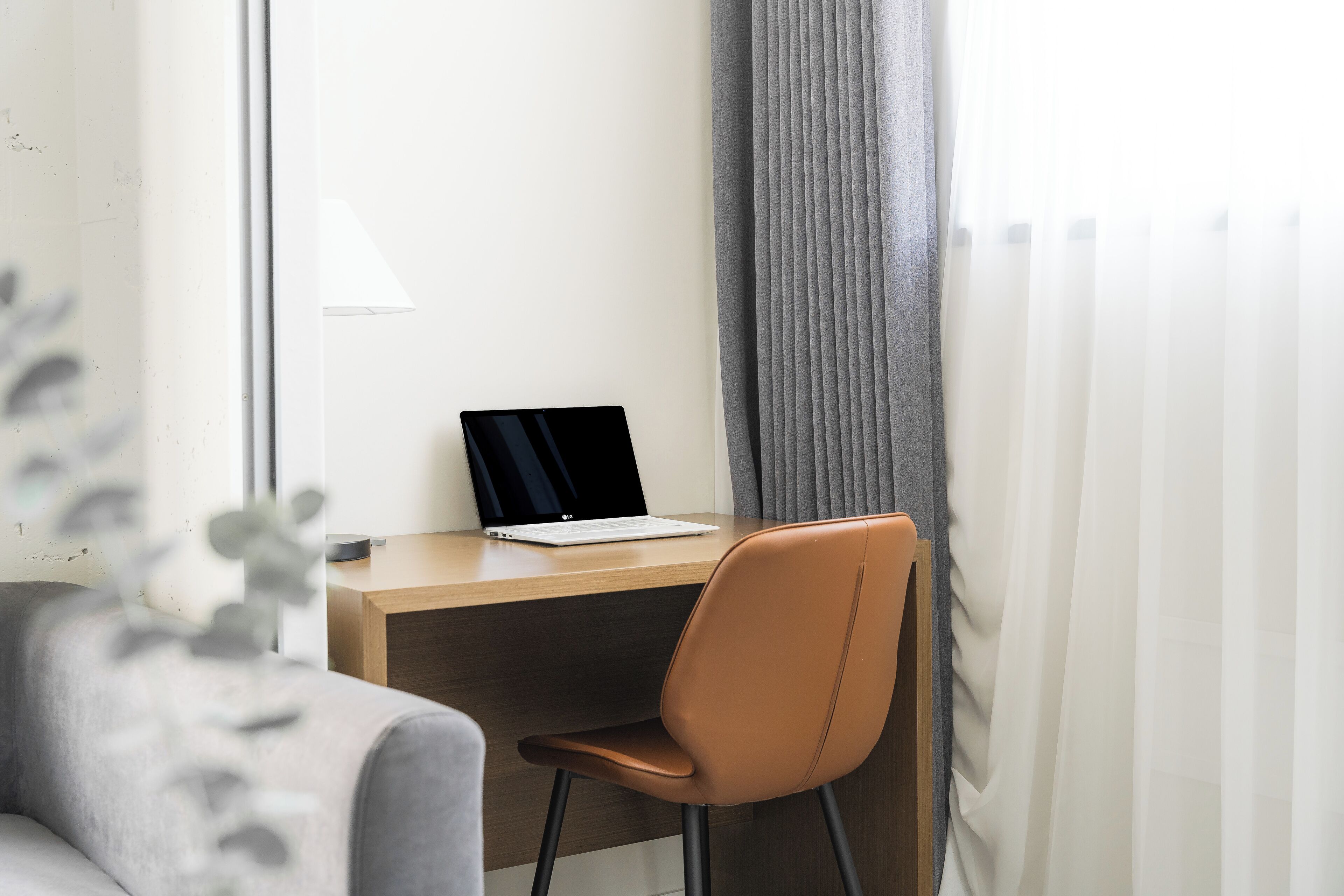 A modern office space with a laptop on a wooden desk, a brown chair, and a grey sofa.