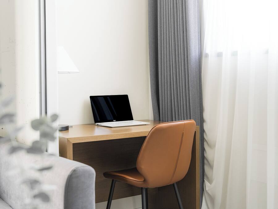 A modern office space with a laptop on a wooden desk, a brown chair, and a grey sofa.