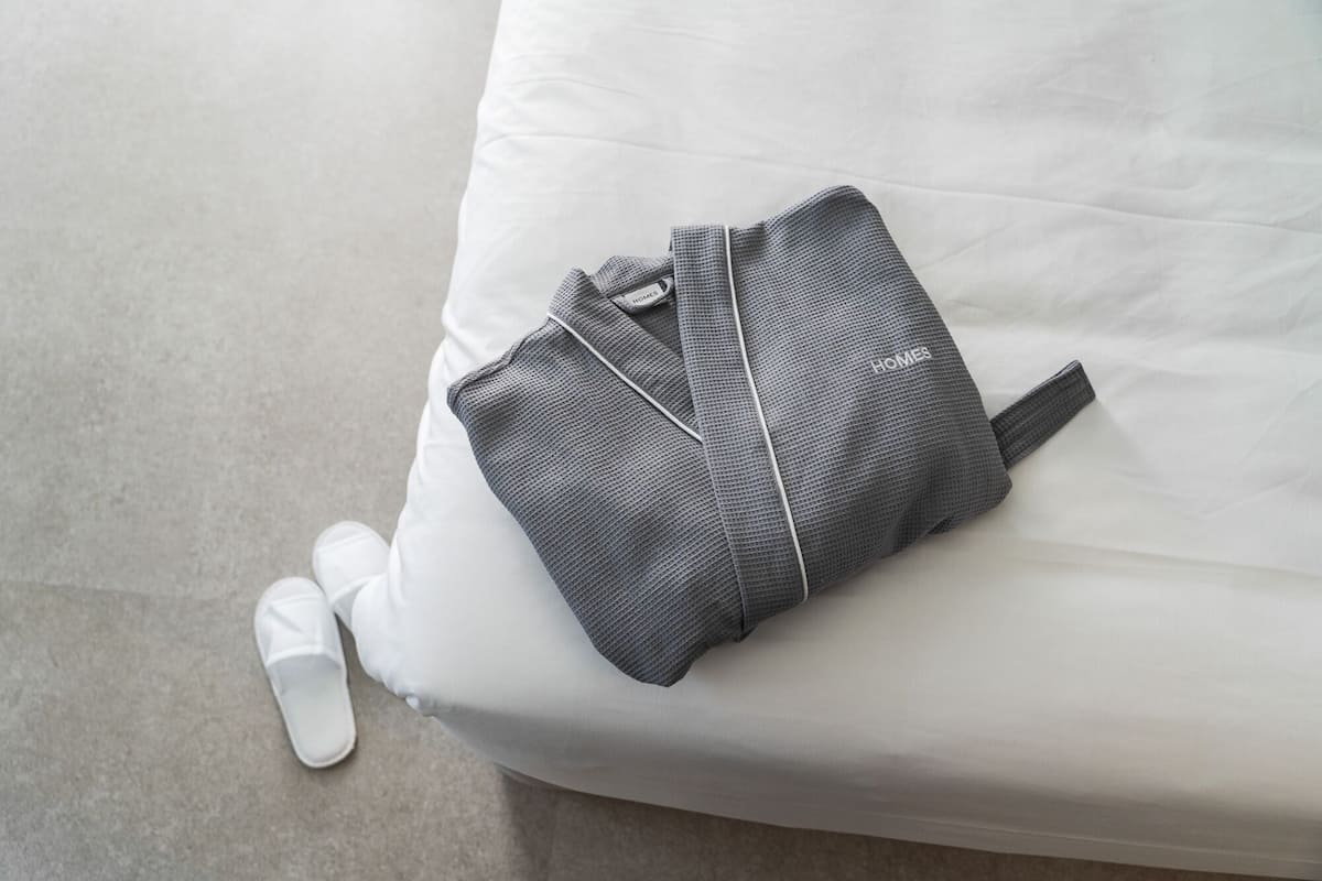 A grey bag on a white bed with a white pillow and a white sock.