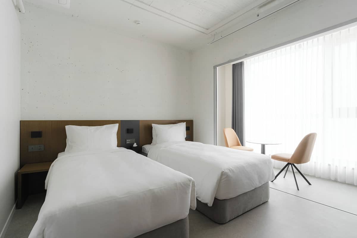 A hotel room with two single beds, a wooden headboard, a small table, a chair, and a large window with blinds.