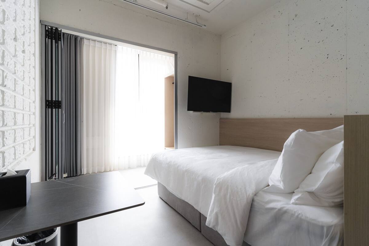 A hotel room with a bed, a desk, a TV mounted on the wall, and a window with blinds.