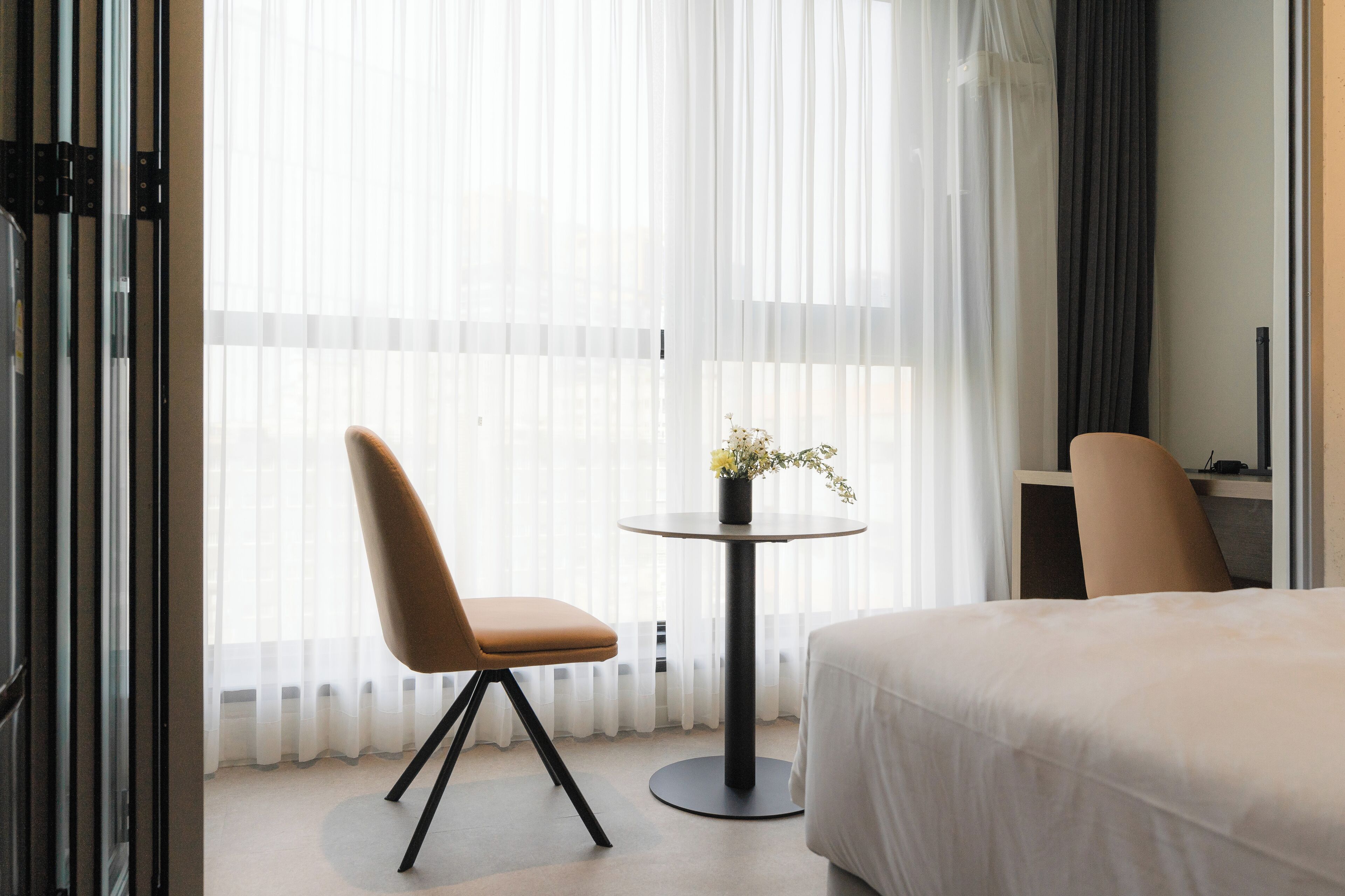 A hotel room with a bed, a chair, a small table with a vase of flowers, and sheer curtains.