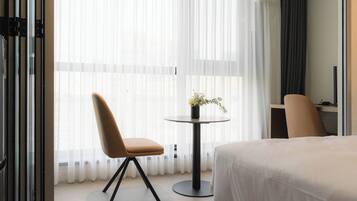 A hotel room with a bed, a chair, a small table with a vase of flowers, and sheer curtains.
