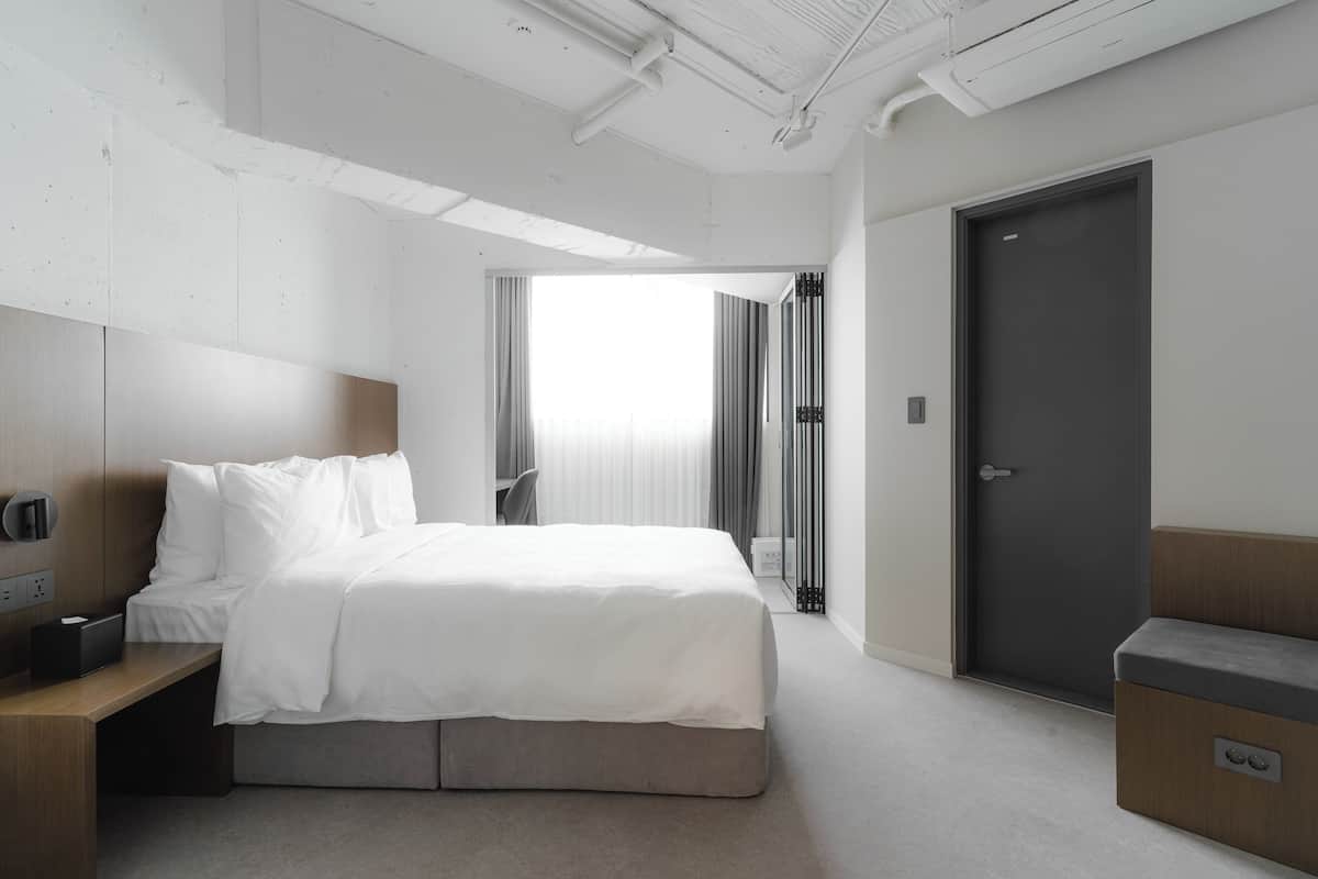 A modern hotel room with a neatly made bed, a bedside table with a lamp, a wardrobe, and a door leading to another room.