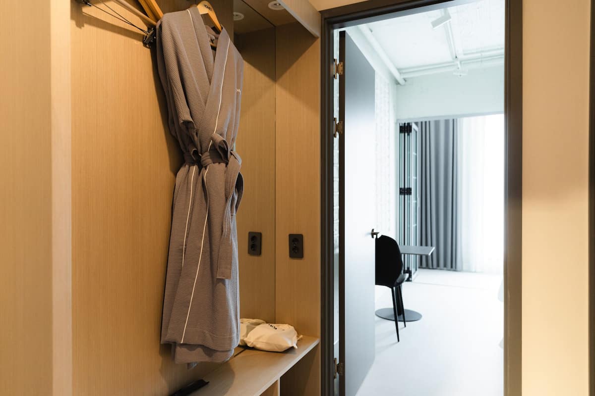 A hotel room with a wooden wardrobe, a robe hanging on the door, a small table with a bag, and a view of a modern bathroom with a chair.