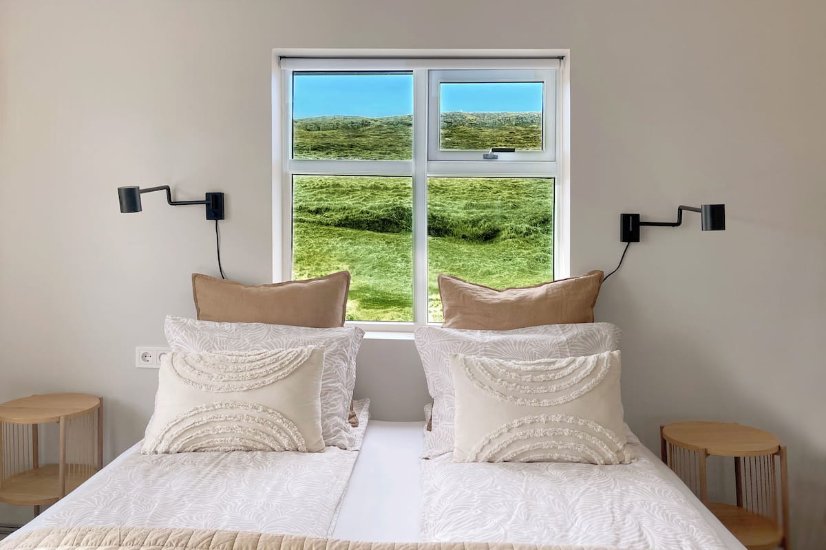 A double bed with a quilted cover, two bedside tables, and a window with a view of a green field.