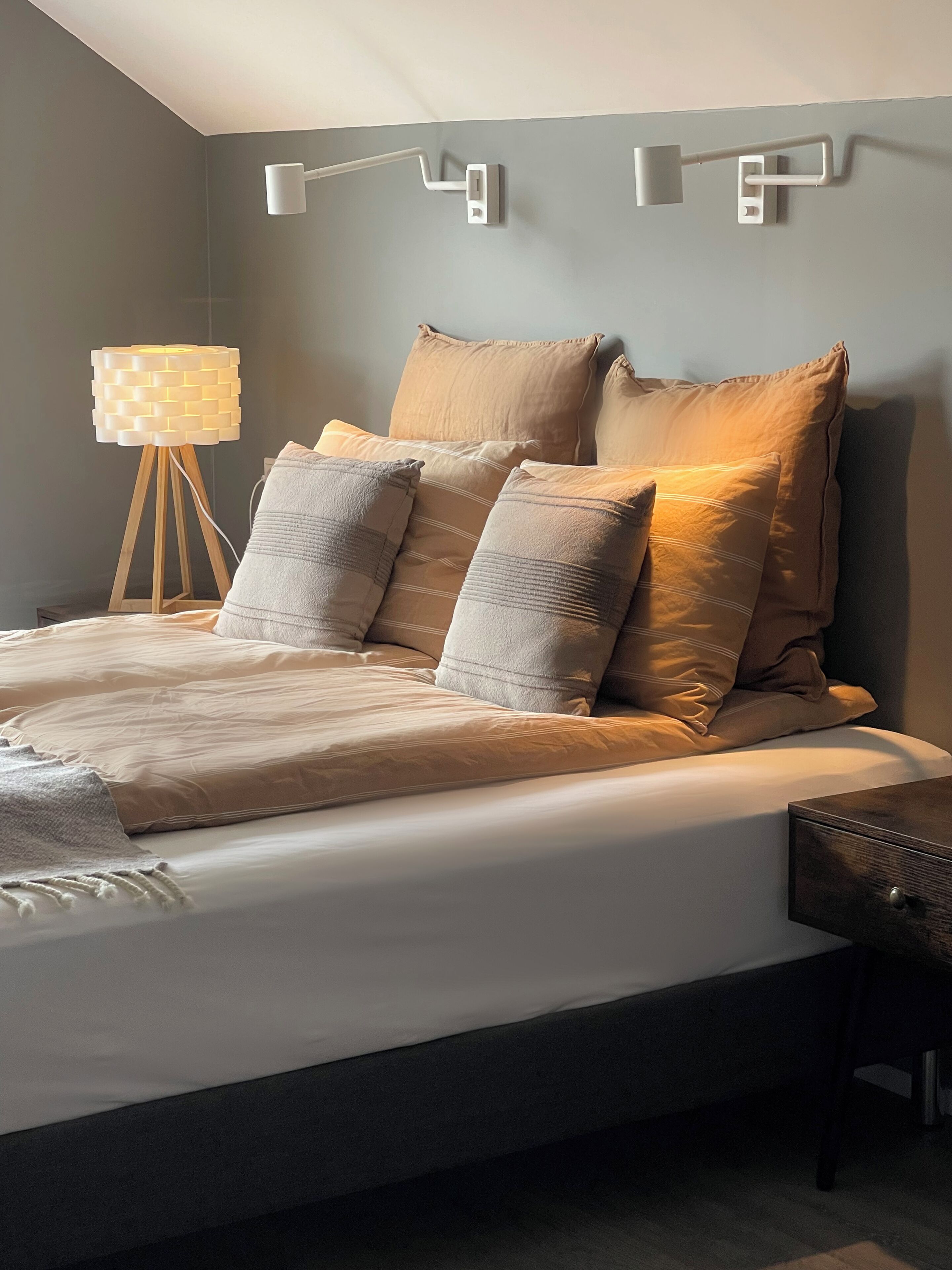 Une chambre moderne avec un grand lit, de nombreux oreillers, une table de chevet et une lampe fixée au mur.