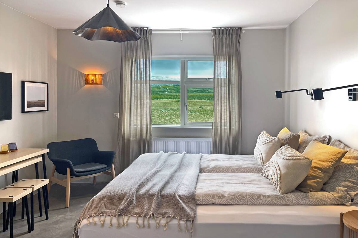 A bedroom with a large bed, a desk, a chair, and a view of a green landscape through the window.