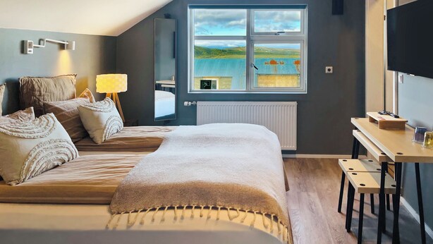 A bedroom with a large bed, a desk with a chair, a TV, a window with a view of a landscape, and a lamp. - Lindarholl Guesthouse (Egilsstaðir)