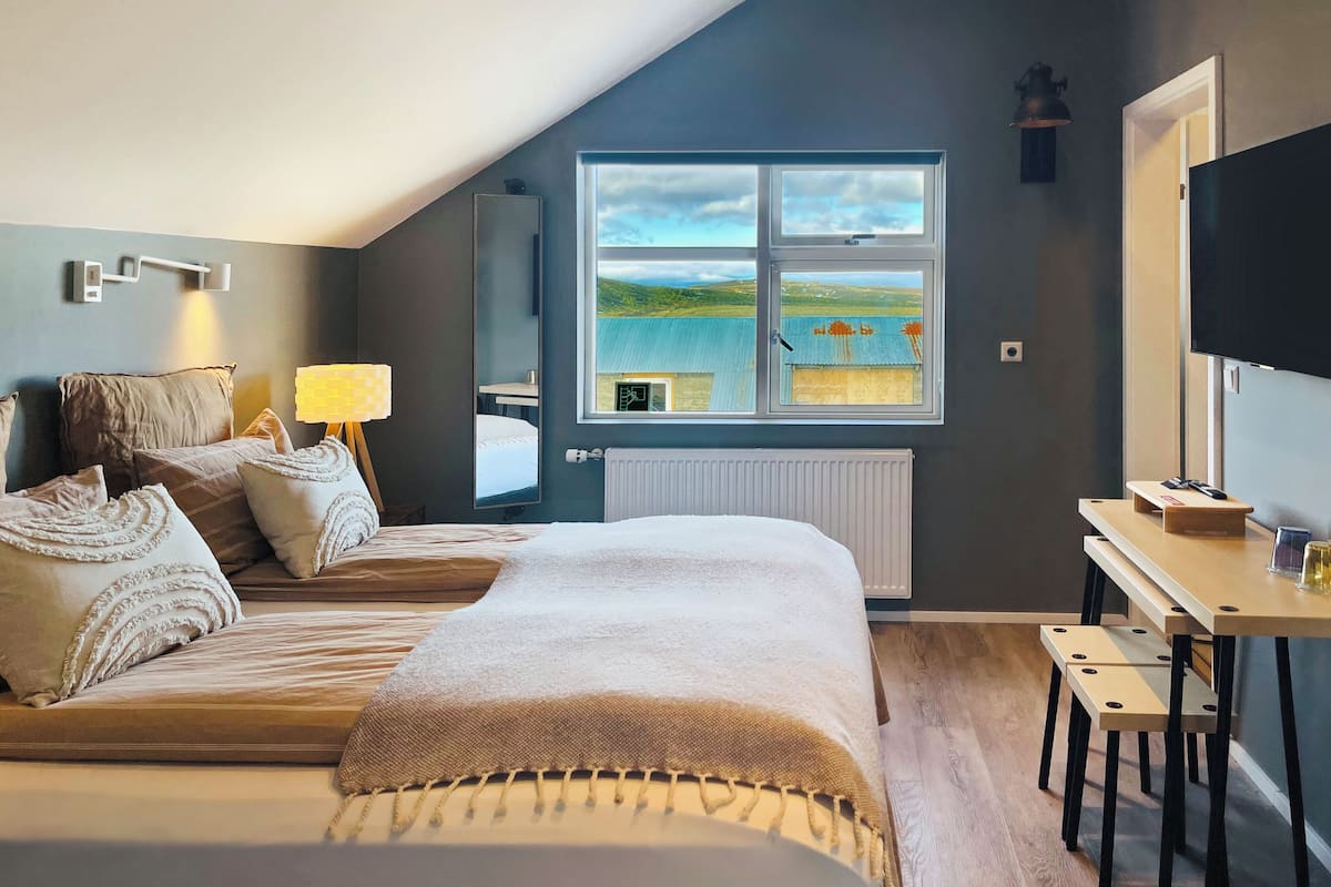 A bedroom with a large bed, a desk with a chair, a TV, a window with a view of a landscape, and a lamp.