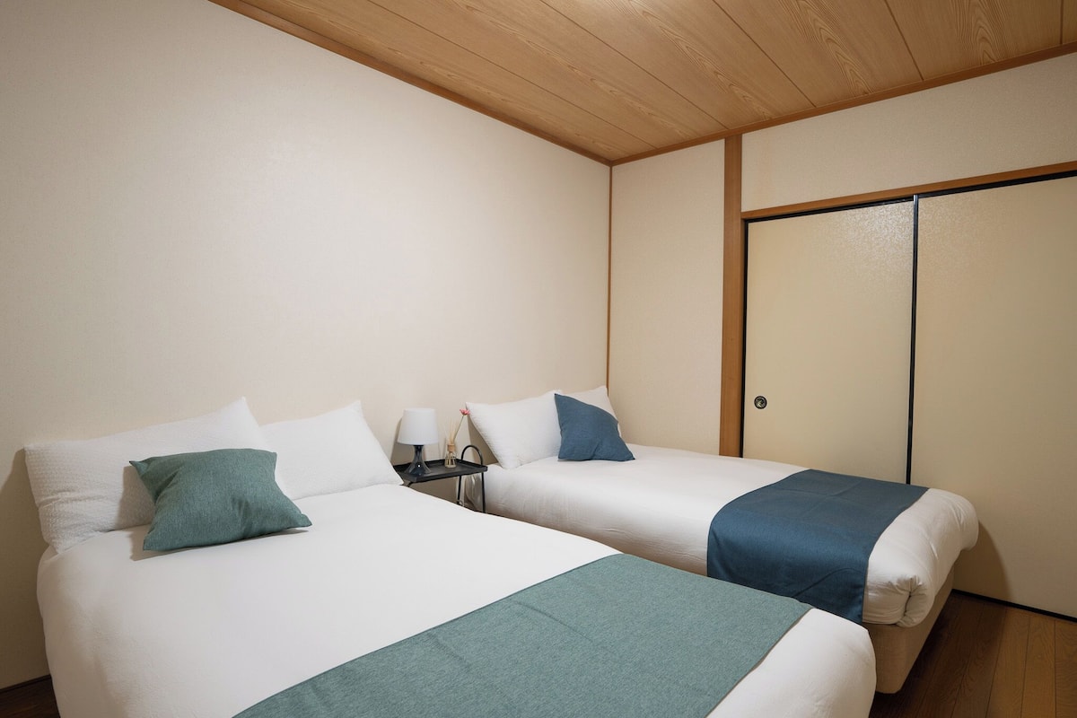 A room with two beds, each with white bedding and teal accents. There is a nightstand with a lamp and a small table with a vase. The room has wooden flooring and a wooden ceiling.