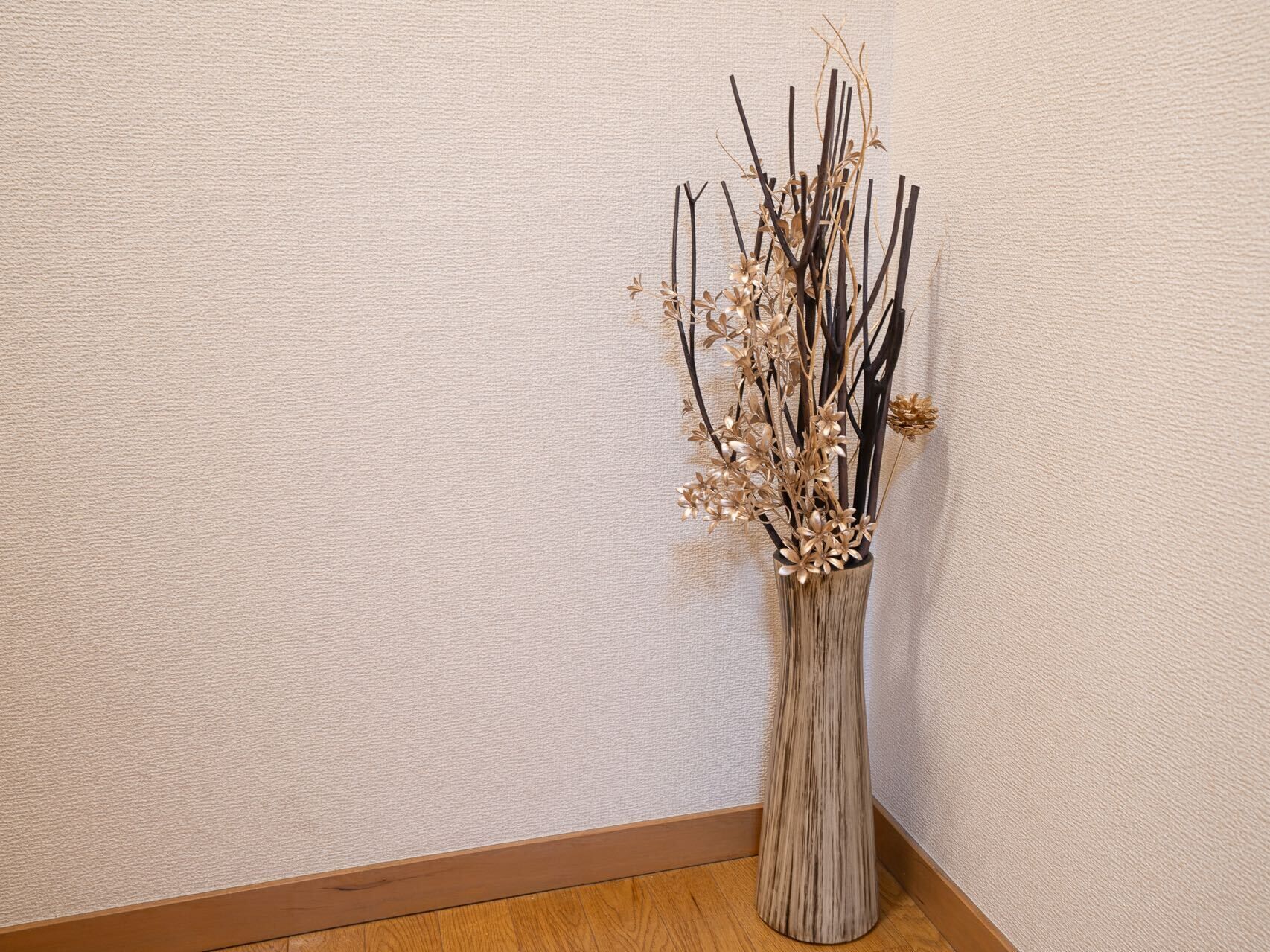 A tall, narrow vase with dried branches and flowers against a textured wall.