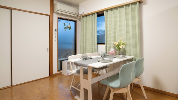 Un coin repas avec une table dressée pour deux personnes, une fenêtre avec des rideaux et une vue sur l’extérieur.