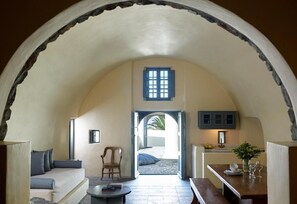 An arched living room with a sofa, a chair, a small table, and a kitchen area visible through an open doorway. - Zannos Melathron Hotel by Omilos Hotels (Santorini)