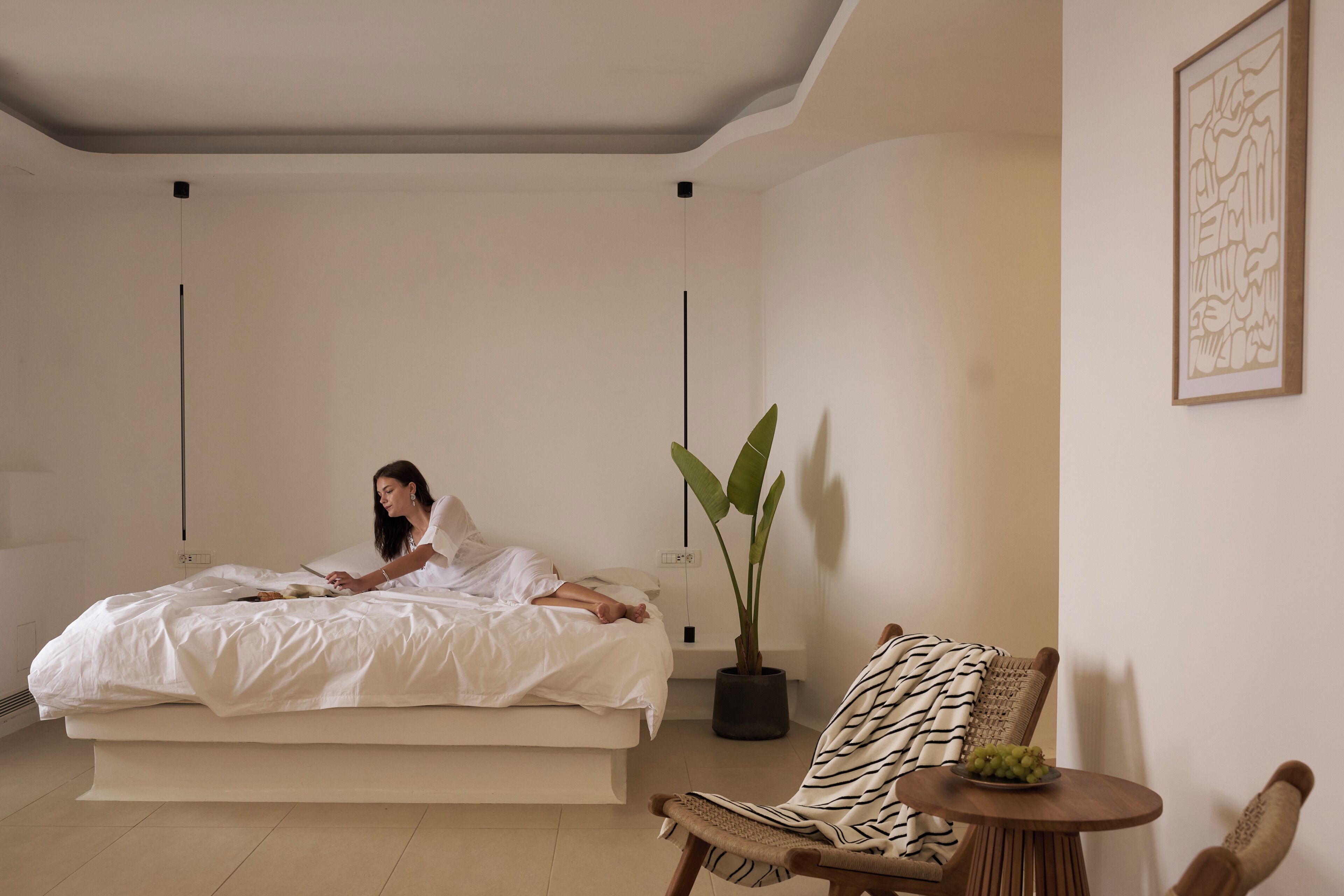 A bedroom with a bed, a potted plant, a chair, and a table with a bowl of fruit.
