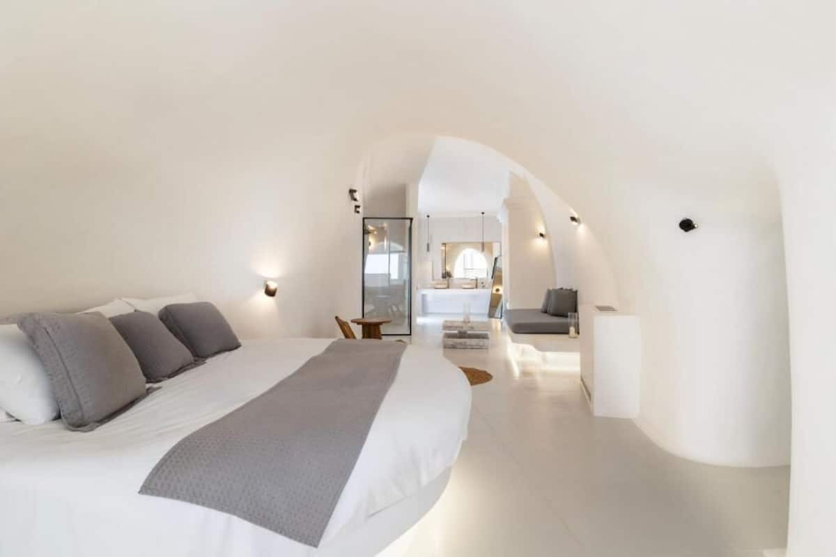 A modern, white-themed bedroom with a large bed, a seating area, and a view into the living space.