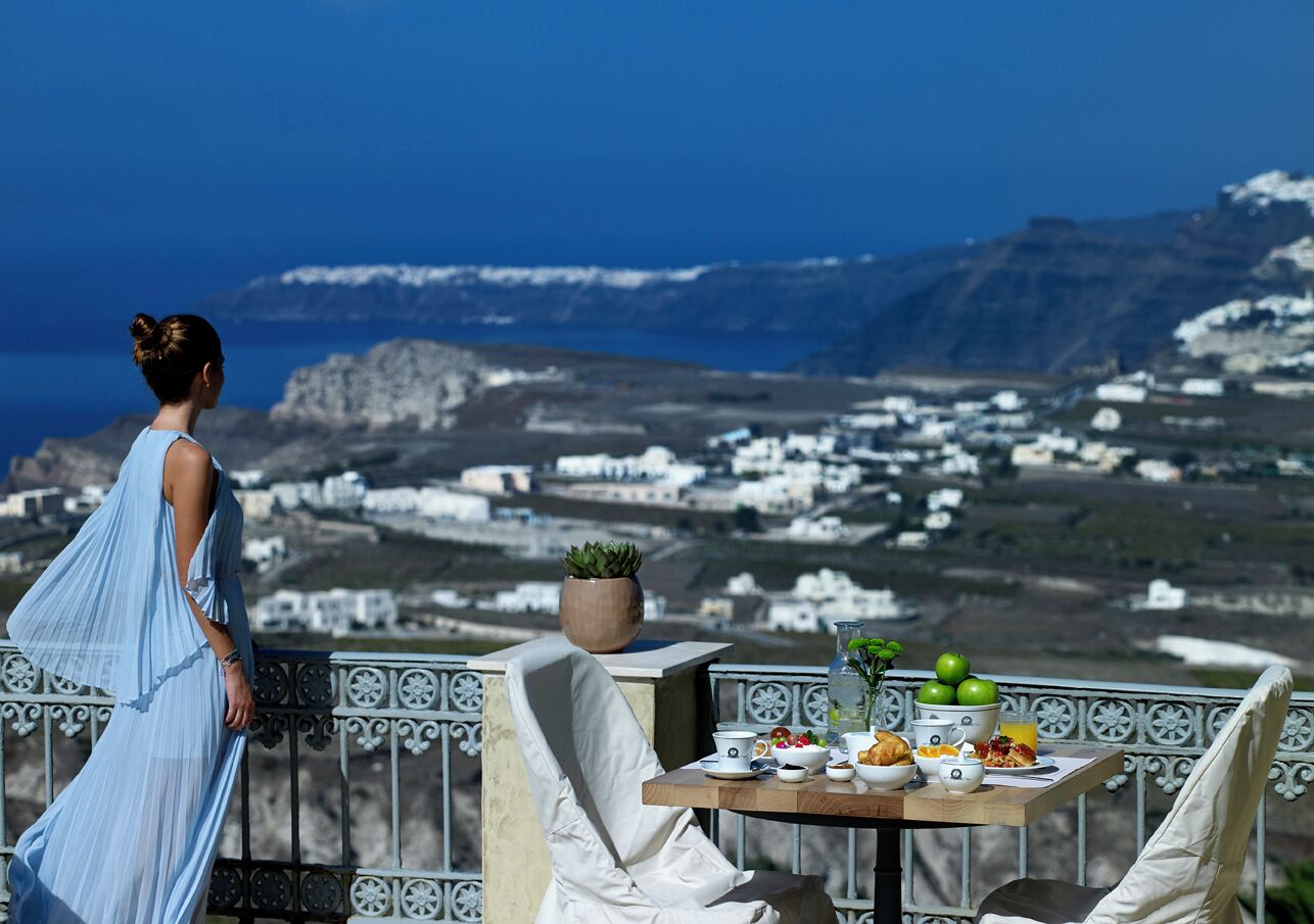 A balcony with a table set for breakfast, overlooking a coastal landscape with white buildings and mountains.