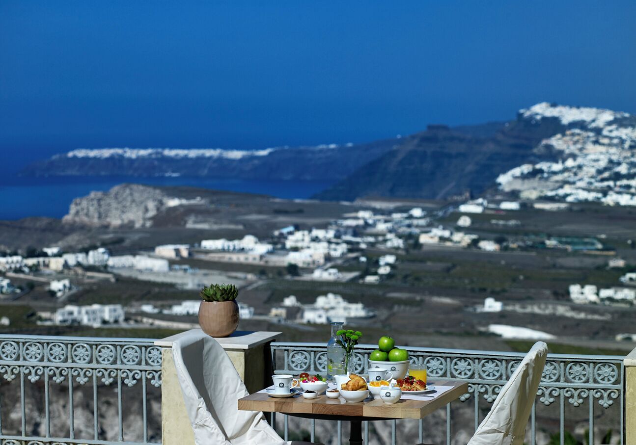 A table with food and drinks, a potted plant, and a view of a coastal village.