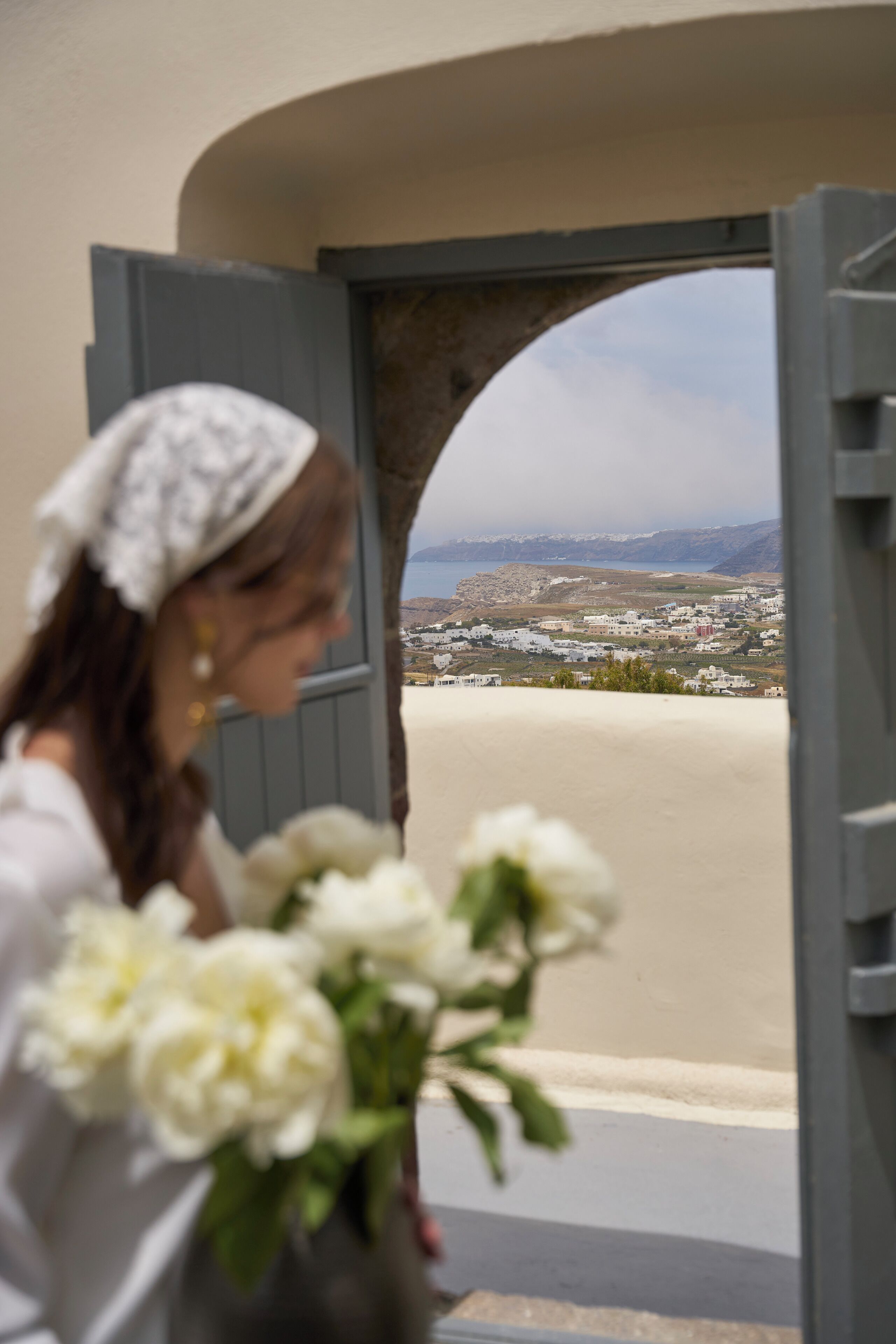 A person holding a bouquet of flowers, standing in front of an open doorway with a view of a coastal landscape.