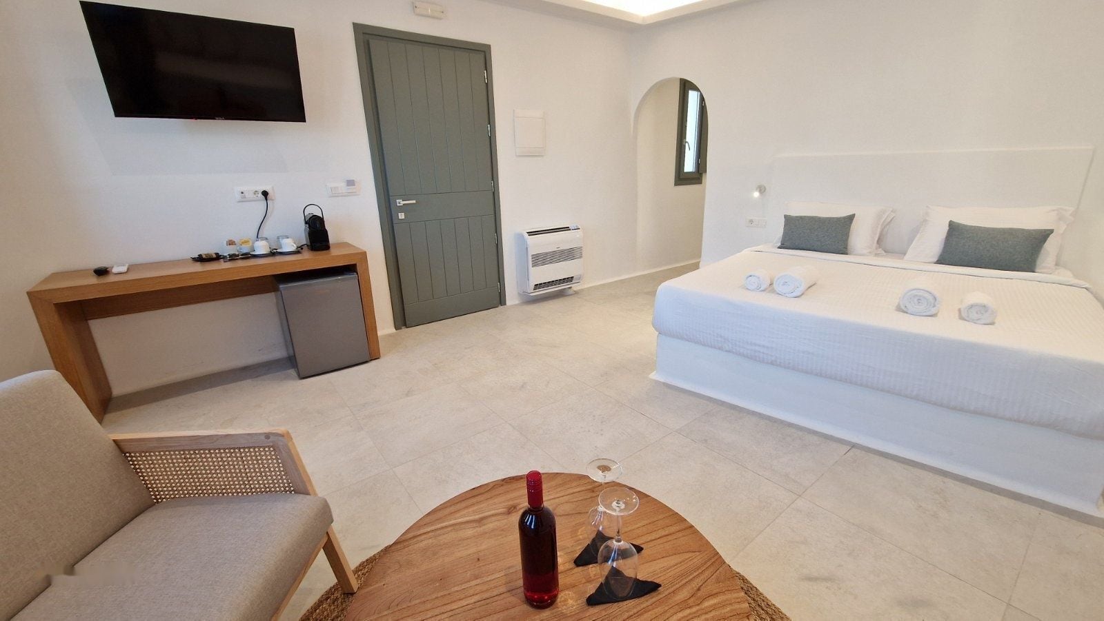 Habitación de hotel con cama, escritorio, televisor y una mesita con una botella y vaso.